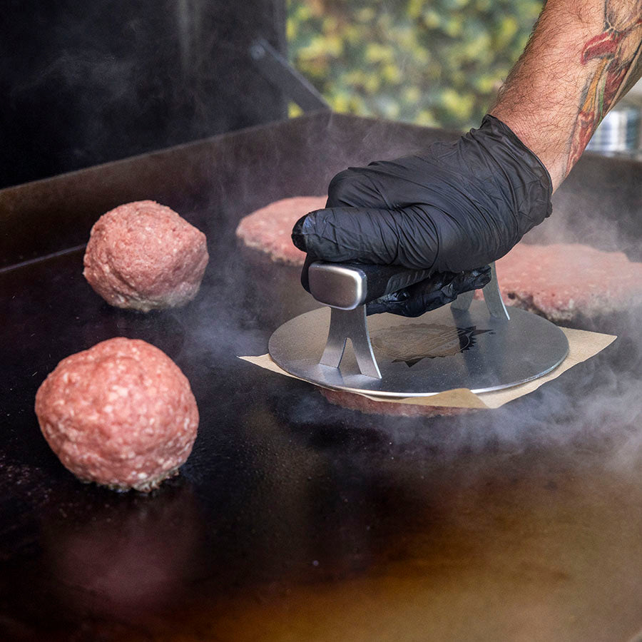 Silver burger press on hot griddle, used by gloved hand.