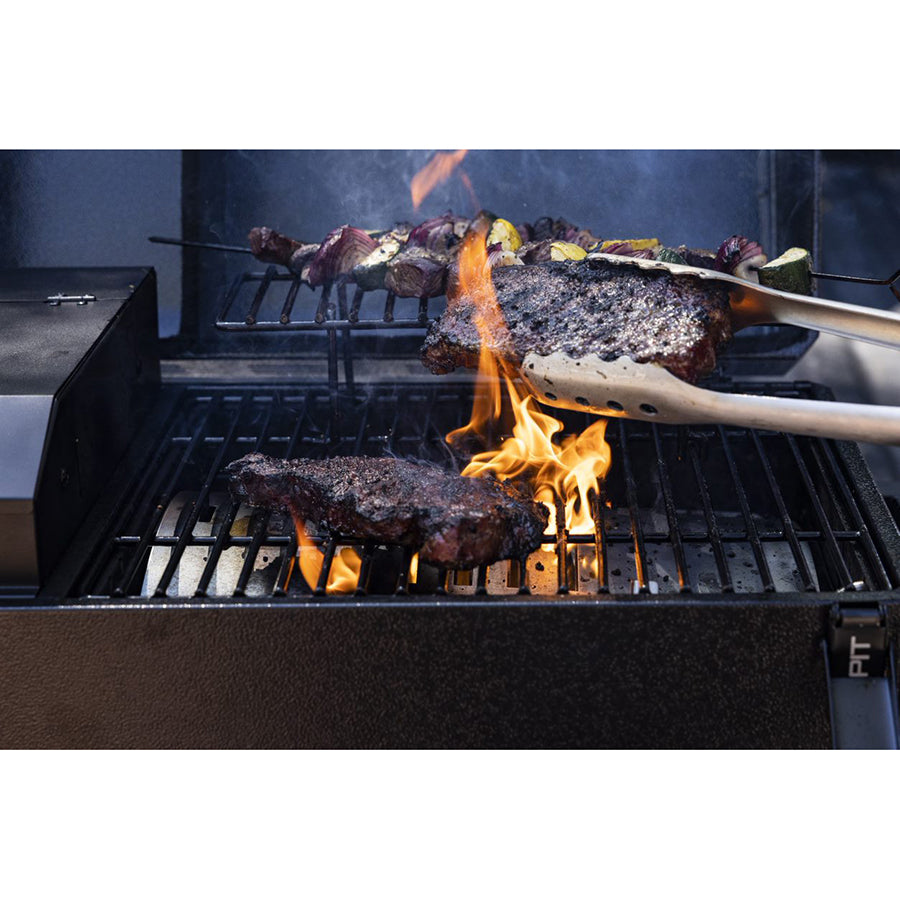 Close-up of steaks and skewered vegetables grilling over open flames on a barbecue grill, with tongs holding one steak.