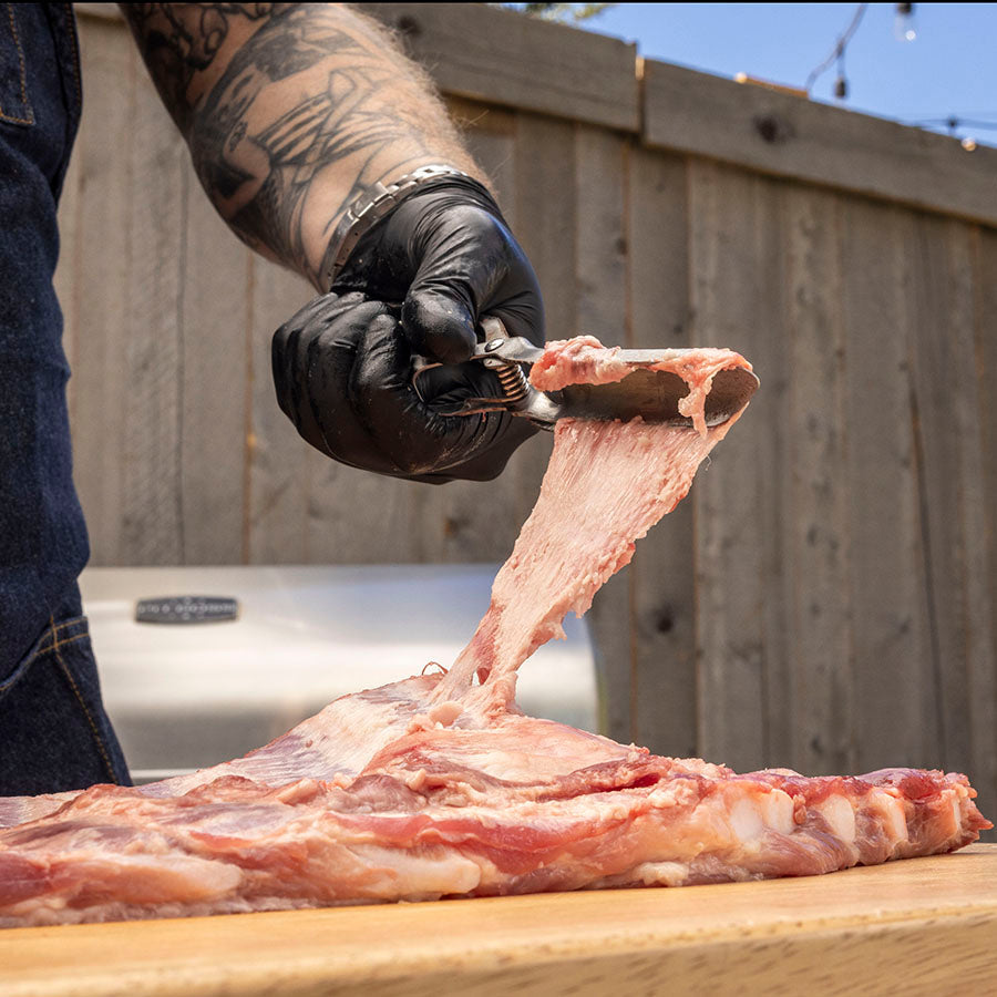 Person wearing black gloves cutting meat with knife on wooden board.