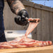 Person wearing black gloves cutting meat with knife on wooden board.