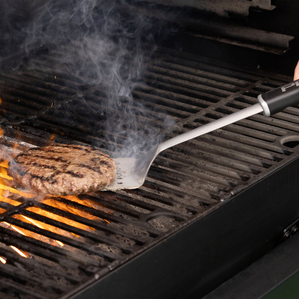Metal spatula flipping a grilled burger patty over open flames on a barbecue grill, with visible smoke rising.