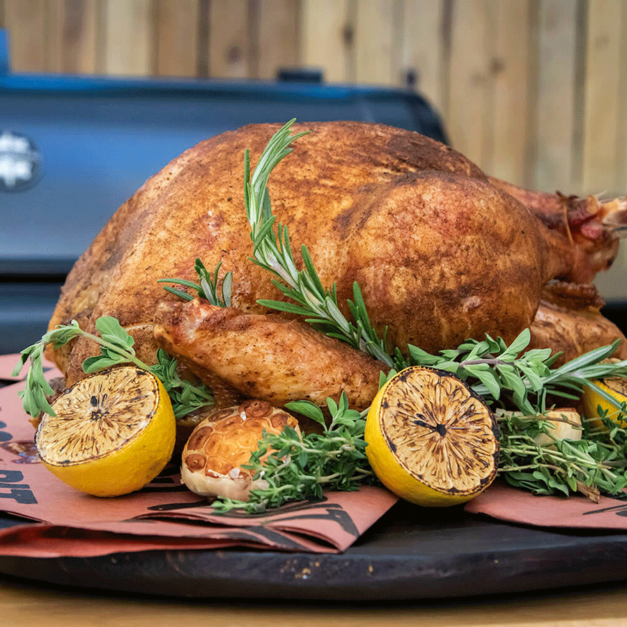 Whole roasted turkey garnished with fresh herbs, grilled lemon halves, and roasted garlic on a wooden platter.