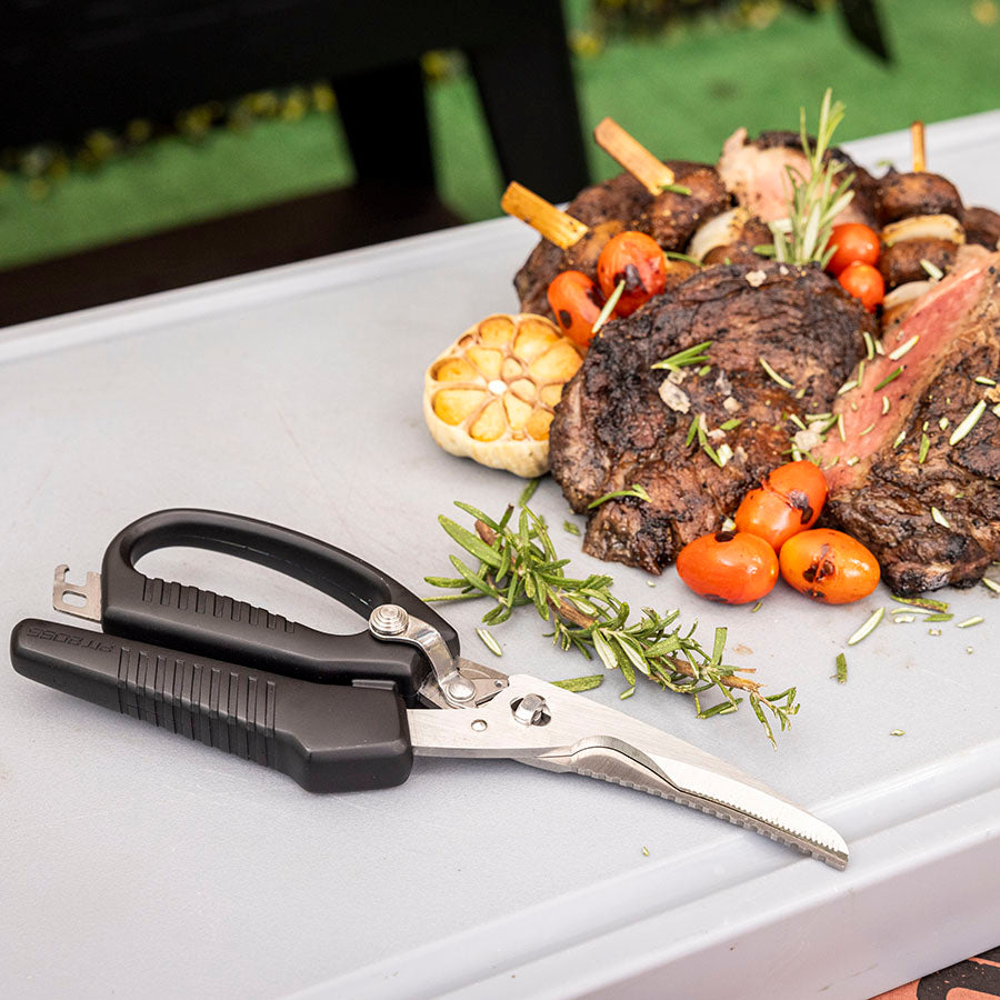 Black-handled kitchen shears with serrated blades, displayed near grilled steak and vegetables.