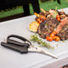 Black-handled kitchen shears with serrated blades, displayed near grilled steak and vegetables.