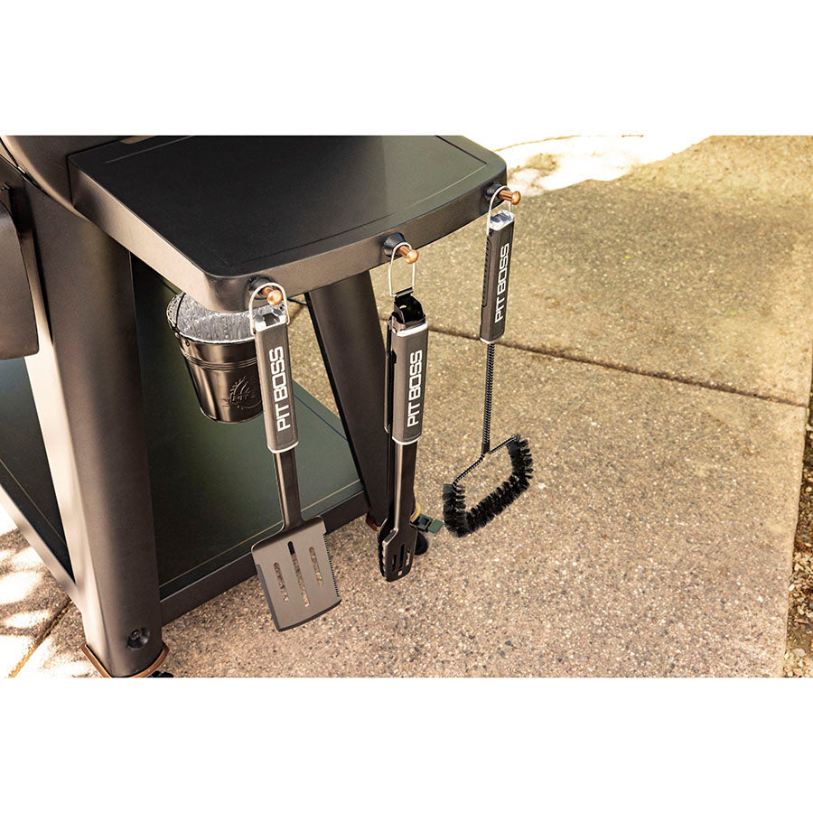"Black Pit Boss BBQ tools hanging from a grill, including a spatula, tongs, and brush."