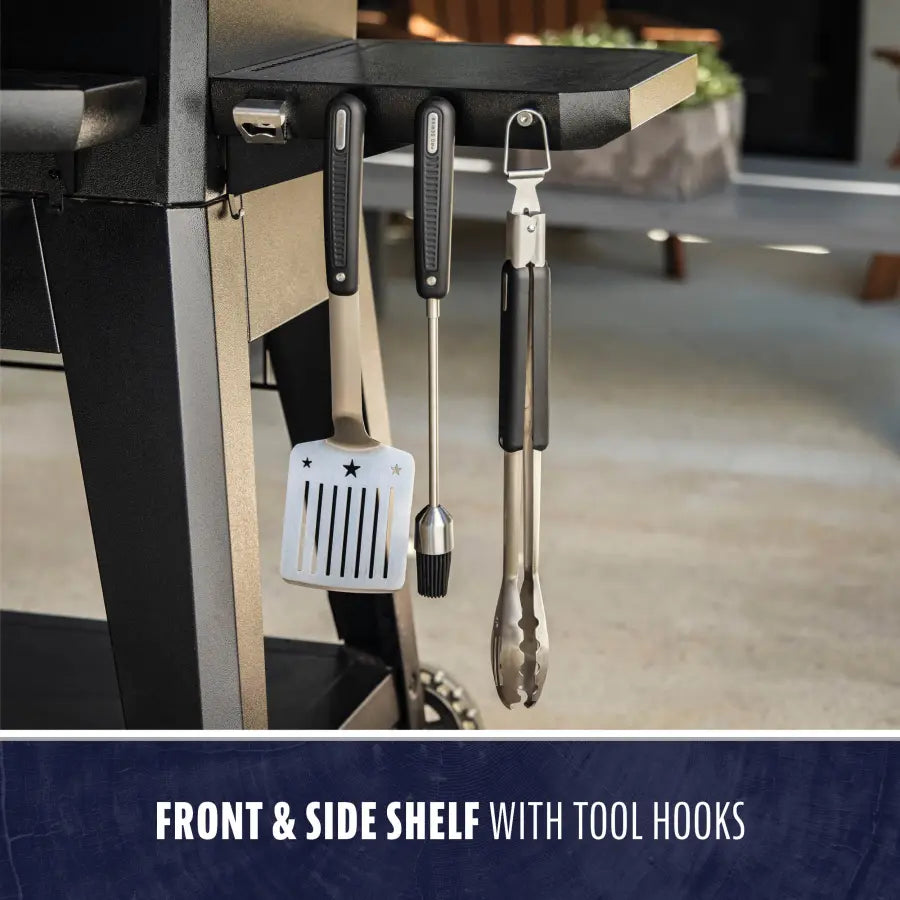 Grill with black front and side shelf, tool hooks holding spatula, brush, and tongs.