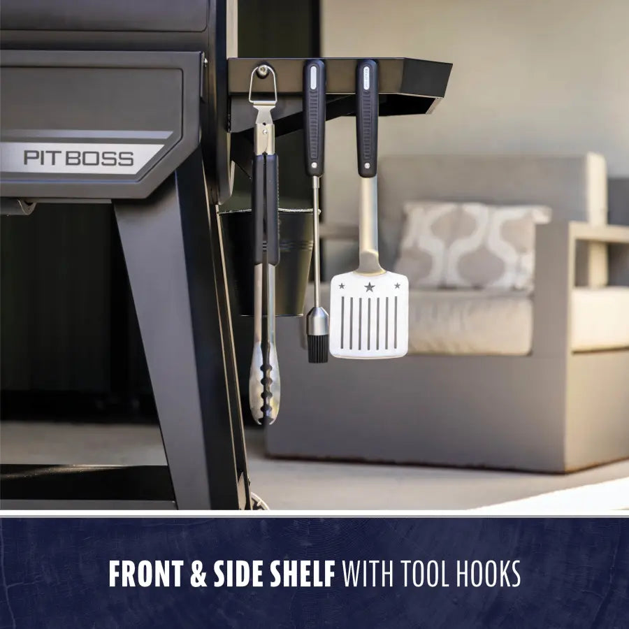 Black Pit Boss grill with front and side shelf, tool hooks holding tongs and spatula.