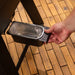 Hand removing a rectangular grill drip tray with a foil liner from a black metal holder on a wooden floor.