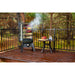 Smoker standing outside on a deck in the trees. Smoker door open. Meats and veggies smoking inside. Mini table displayed next to the smoker with meat and water glasses on it.
