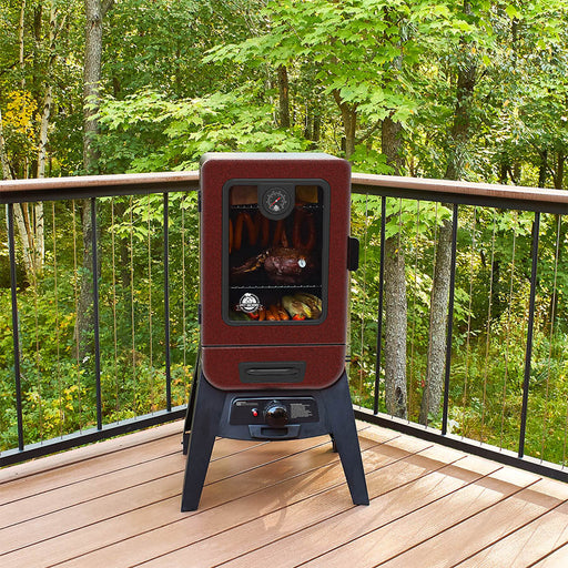 Vertical smoker outside o a deck in the trees. Meats and veggies cooking inside the smoker.