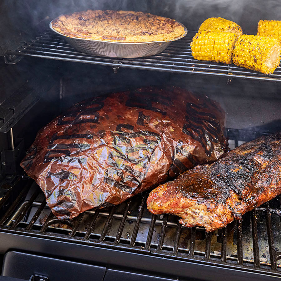Close up of butcher paper wrapped around meat inside grill next to other meats and veggies on both grilling racks.