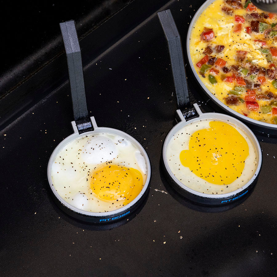 Pit Boss 2 Pack Silicone Egg Molds, grey and blue with pit boss logo, 4 inch diameter each, eggs cooking on griddle top in egg rings