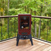 Vertical smoker outside o a deck in the trees. Meats and veggies cooking inside the smoker.