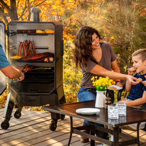lifestyle_4, Black smoker with small Pit boss logo on transparent door and engraved "Pro Series" on front. Family smoking food by a table/