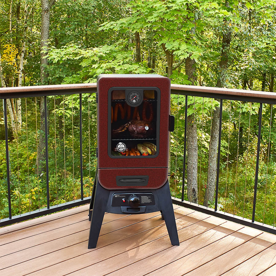 Vertical smoker outside o a deck in the trees. Meats and veggies cooking inside the smoker.