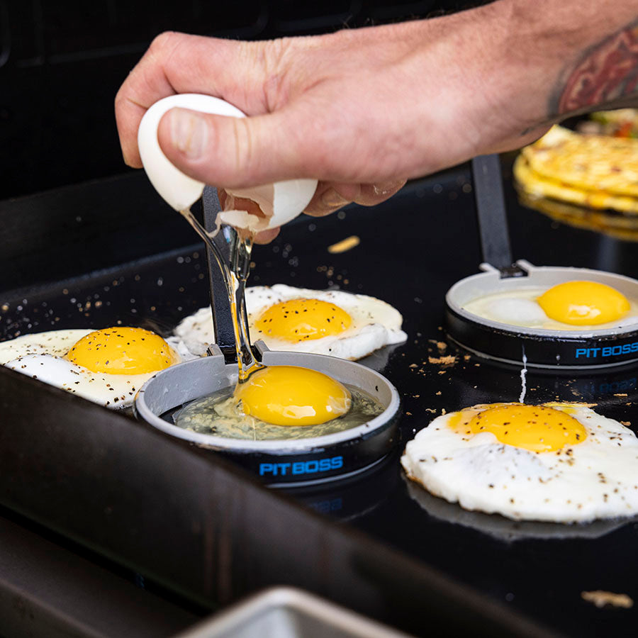 Pit Boss 2 Pack Silicone Egg Molds, grey and blue with pit boss logo, 4 inch diameter each, cracking raw eggs into molds on griddle top