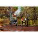 Smoker standing outside on a deck in the trees. Smoker door open. Meats and veggies smoking inside. Mini table displayed next to the smoker with meat and water glasses on it.
