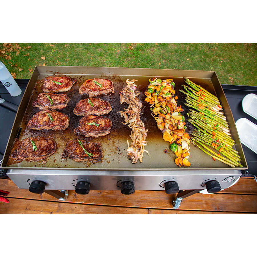 Silver flat-top grill cooking steaks, onions, vegetables, and asparagus.