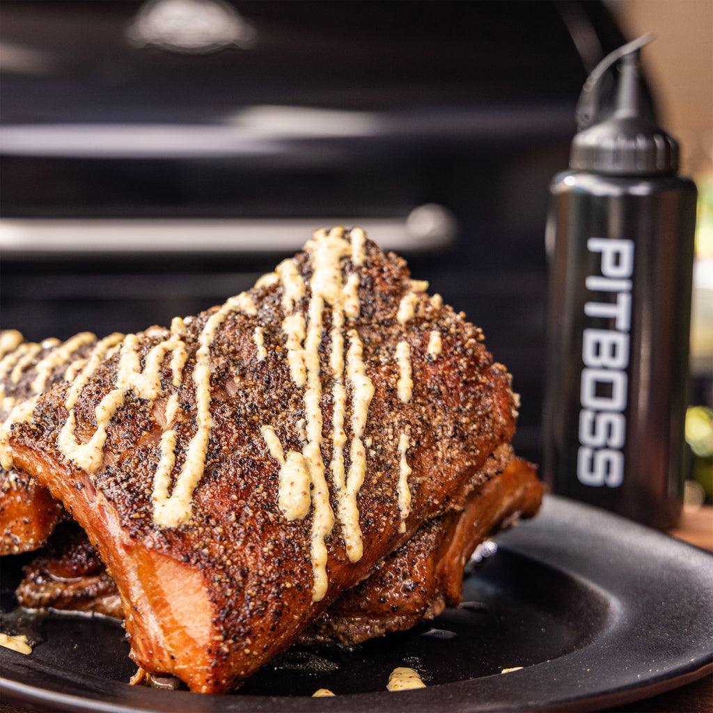 Grilled ribs with pepper seasoning and creamy sauce drizzle on a black plate, next to a PIT BOSS sauce bottle.