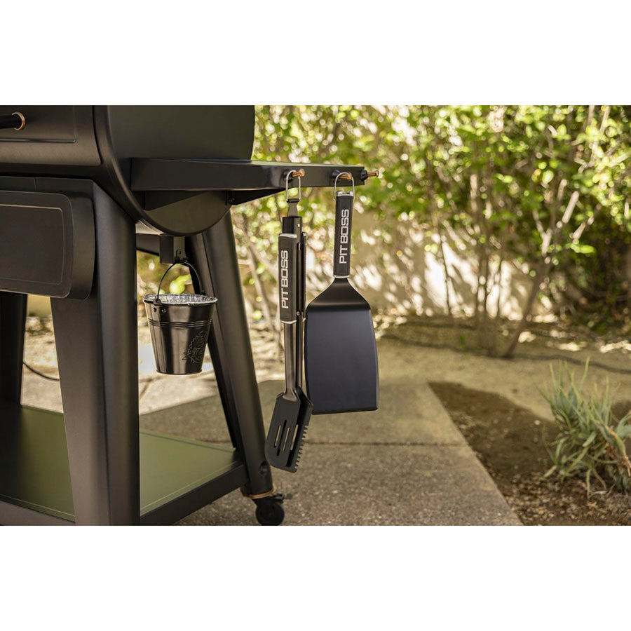 Black Pit Boss grill accessories hanging on a barbecue with a bucket below.