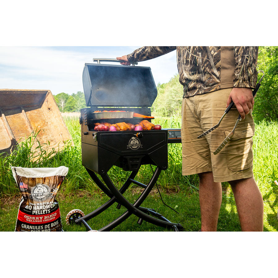 Portable black pellet grill with open lid, grilling meat and vegetables outdoors. Bag of hardwood pellets beside grill.