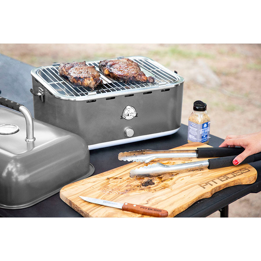 Portable tabletop grill with steaks cooking, metal tongs, knife, and seasoning on a wooden cutting board labeled "Pit Boss."