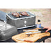 Portable tabletop grill with steaks cooking, metal tongs, knife, and seasoning on a wooden cutting board labeled "Pit Boss."