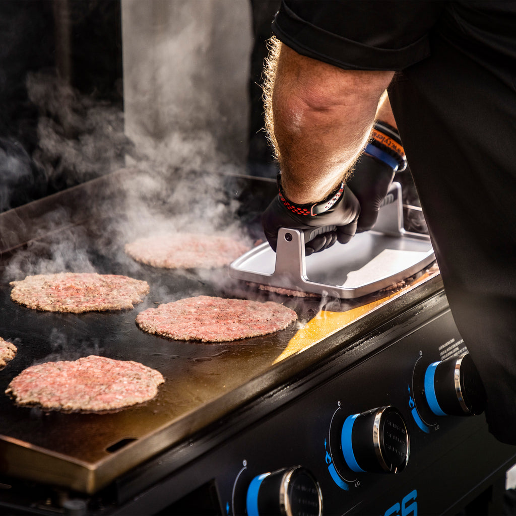 Person pressing burger patties on a flat top griddle with a metal press; steam rising, blue control knobs visible.
