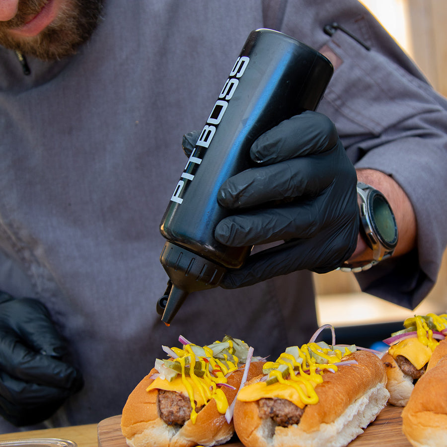 Black Pit Boss squeeze bottle dispensing mustard onto hot dogs.
