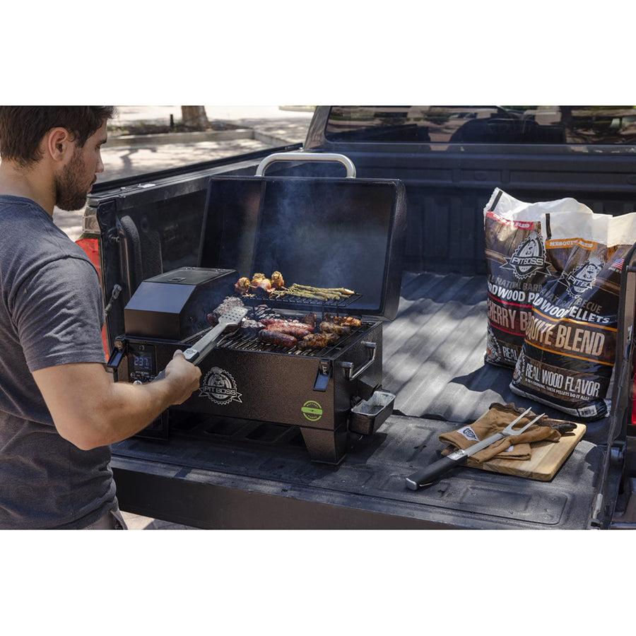 Portable pellet grill on a truck bed, grilling meat and veggies. Includes wood pellets and grilling tools nearby.