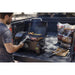Portable pellet grill on a truck bed, grilling meat and veggies. Includes wood pellets and grilling tools nearby.