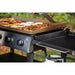 Black gas grill with flat cooking surface, food cooking, outdoor setting.