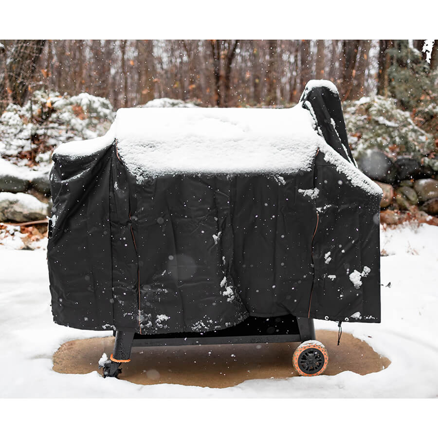 Black outdoor grill cover with wheels, snow-covered in winter setting.