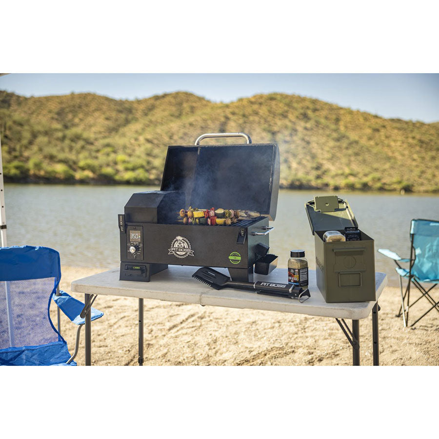 Portable black pellet grill with digital controls, open lid, grilling skewers, on a white table outdoors by a lake.