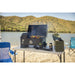 Portable black pellet grill with digital controls, open lid, grilling skewers, on a white table outdoors by a lake.