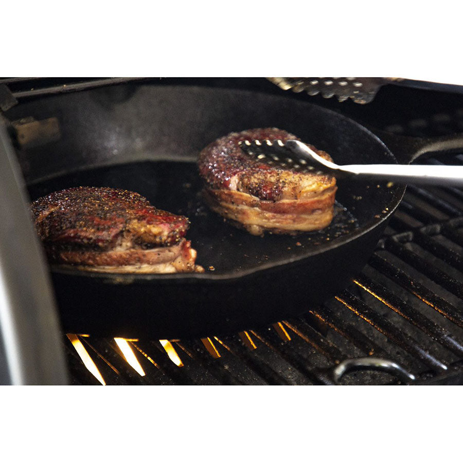 Black cast iron skillet with bacon-wrapped steaks cooking on grill.