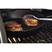 Black cast iron skillet with bacon-wrapped steaks cooking on grill.