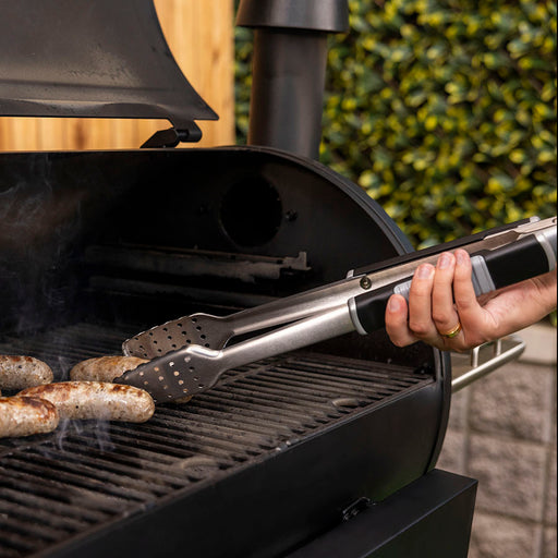 Silver tongs flipping sausages on black grill with smoke.