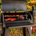 Black outdoor pellet grill with open lid, showing ribs, brisket, vegetables, and smoke inside. Sturdy legs and side shelf.