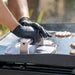 Stainless steel burger press on a griddle, used with black gloves to flatten patties while cooking.