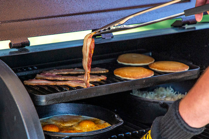 Grill cooking bacon, pancakes, and eggs with tongs.
