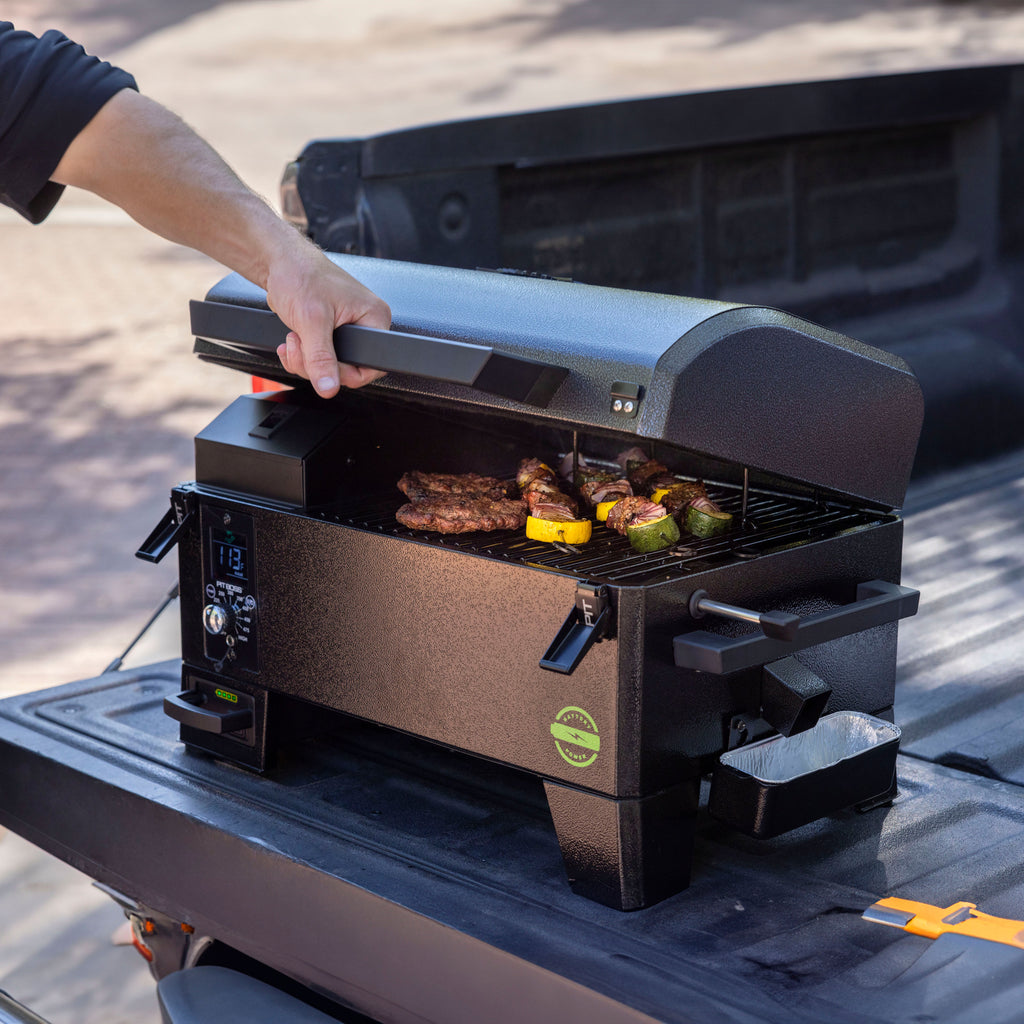 Compact black portable pellet grill with digital controls, open lid, grilling meat and veggies, set on a truck bed.