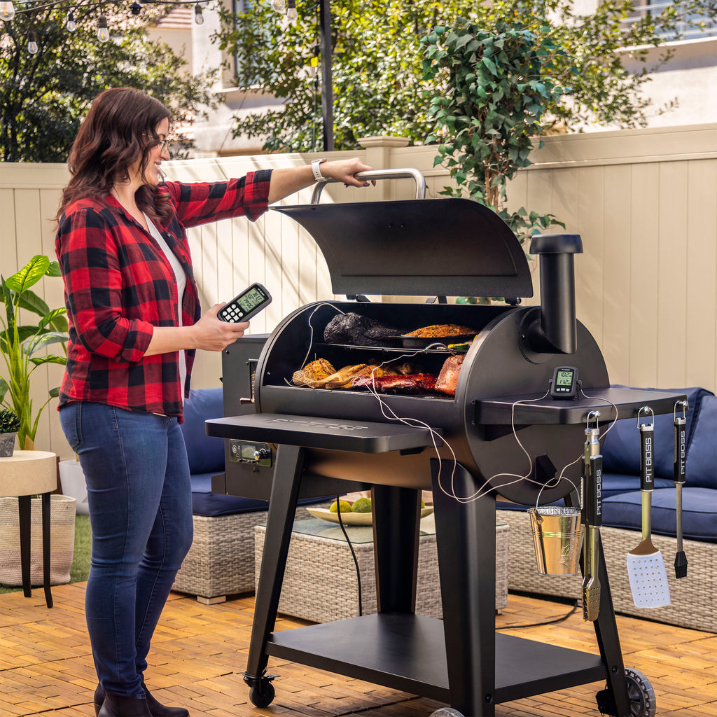 Woman using a black pellet grill with digital controls and meat probes, grilling assorted meats outdoors. Grill tools hang...