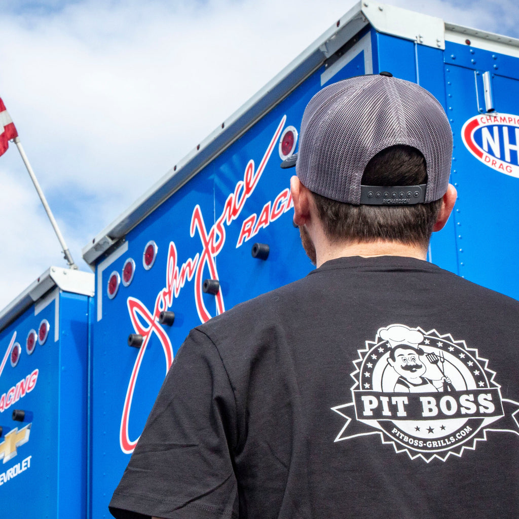 Black Pit Boss t-shirt with logo, worn by person near NHRA trailer.