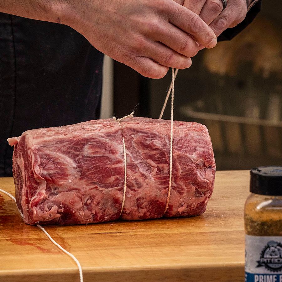 Raw beef tied with string on wooden surface, seasoning bottle nearby.