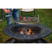 Round outdoor fire pit with a metal grill grate, wood burning inside, and steaks cooking on top.