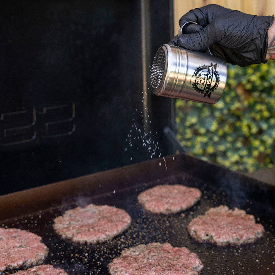 Silver Pit Boss seasoning shaker being used to season burger patties on a grill.