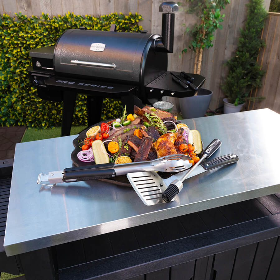 Black Pit Boss Pro Series pellet grill with accessories and cooked food on stainless steel table.