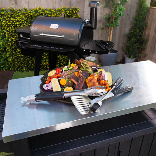 Black Pit Boss Pro Series pellet grill with accessories and cooked food on stainless steel table.