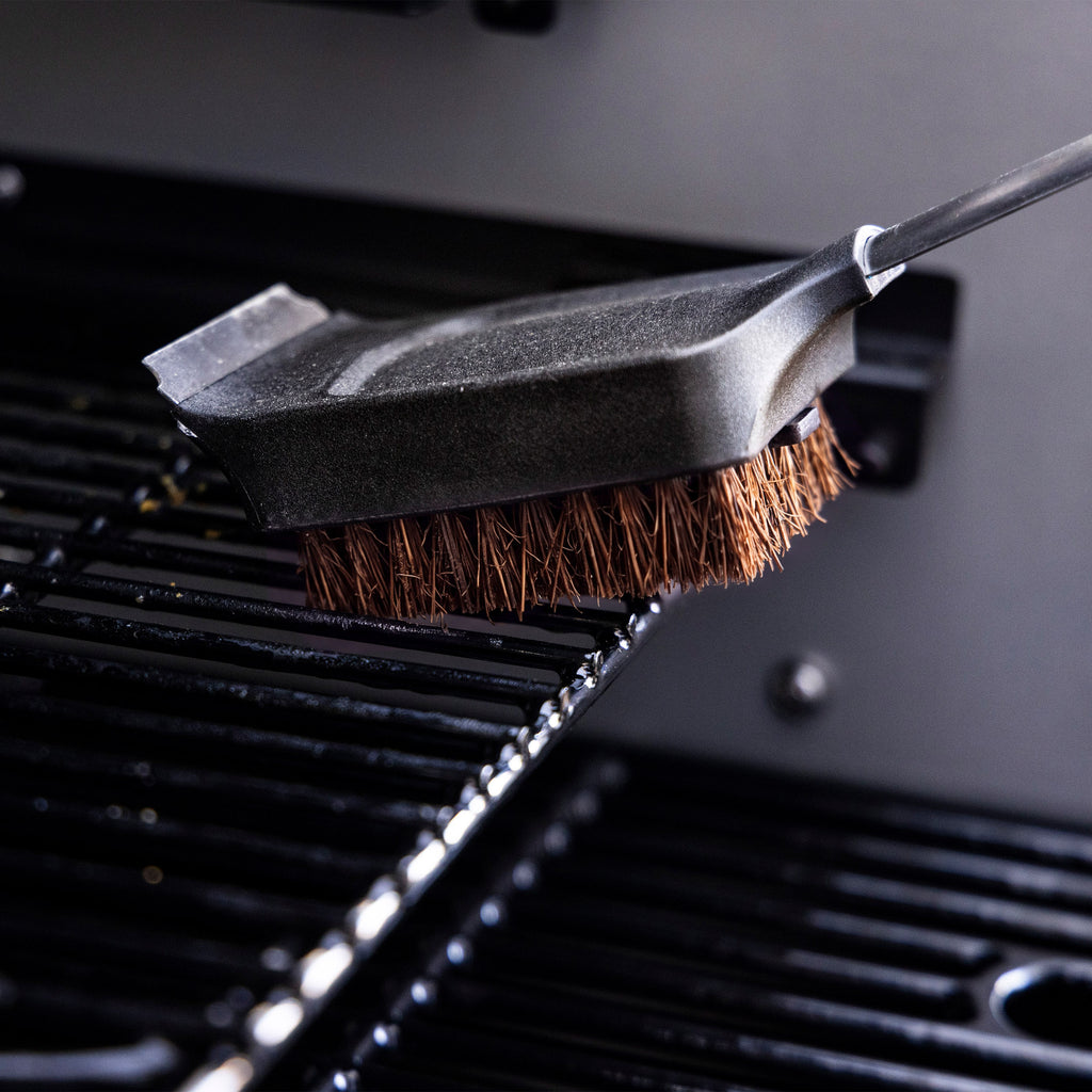 Grill cleaning brush with stiff bristles and metal scraper, shown scrubbing a black grill grate.